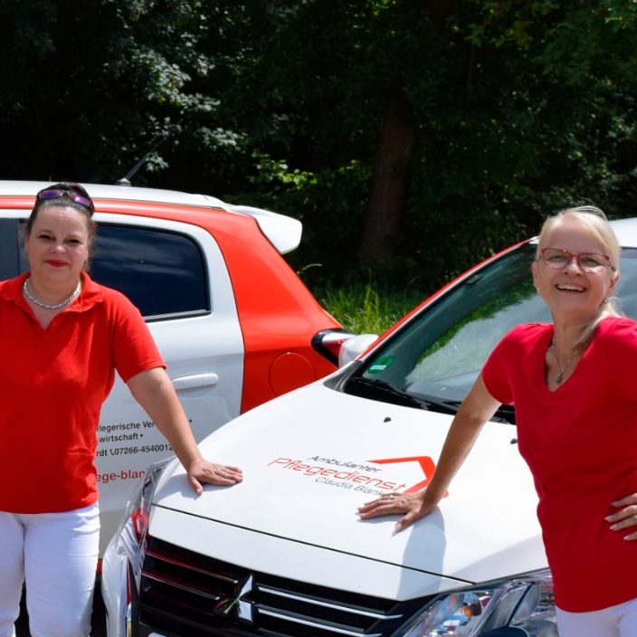 Zwei Frauen in roten T-Shirts stehen vor zwei weißen Autos mit Logo des Pflegedienstes.